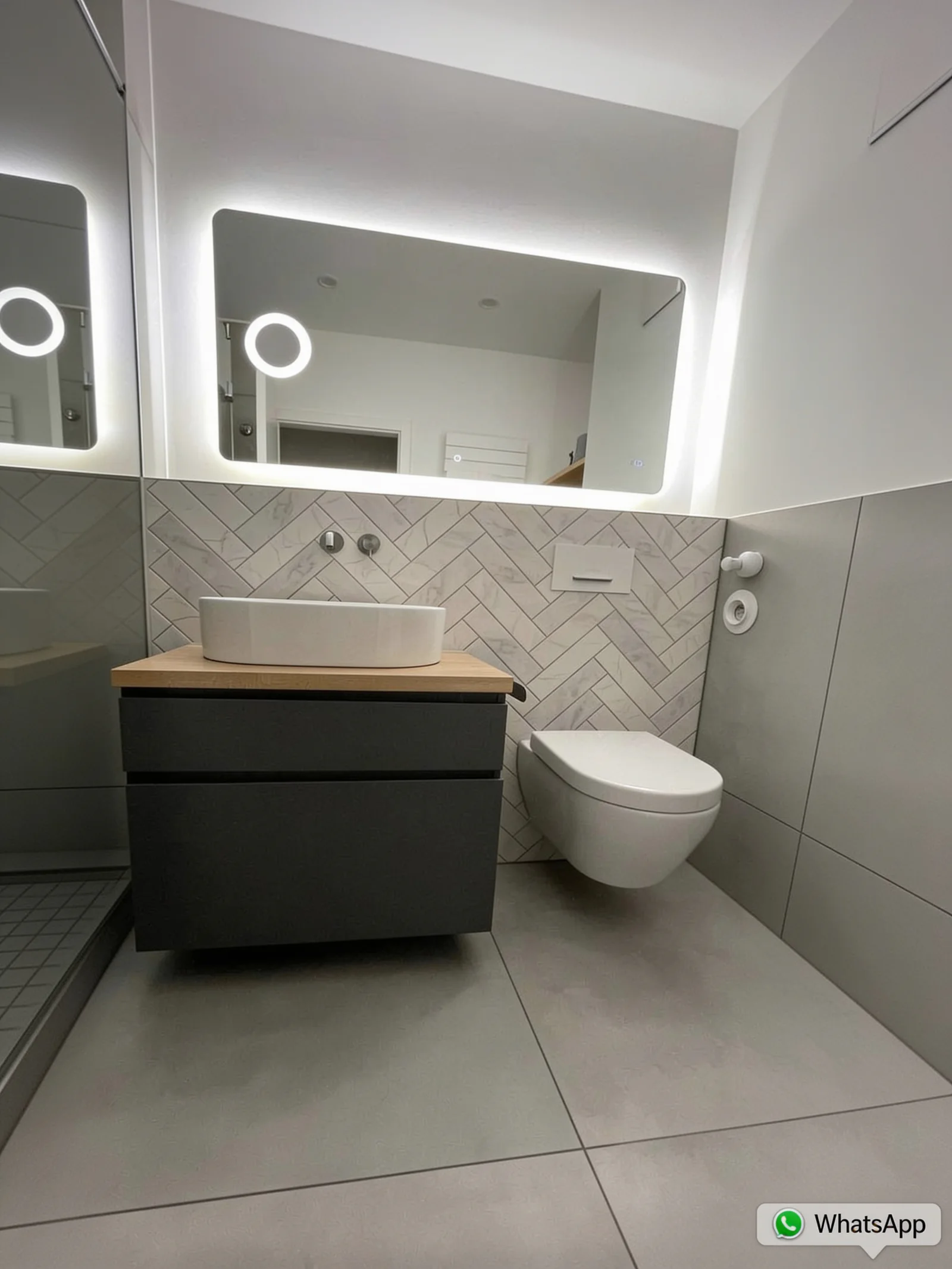 Modernisiertes Bad mit grauen Großformatfliesen, dunklem Waschtischunterschrank, Aufsatzbecken und beleuchtetem Spiegel
