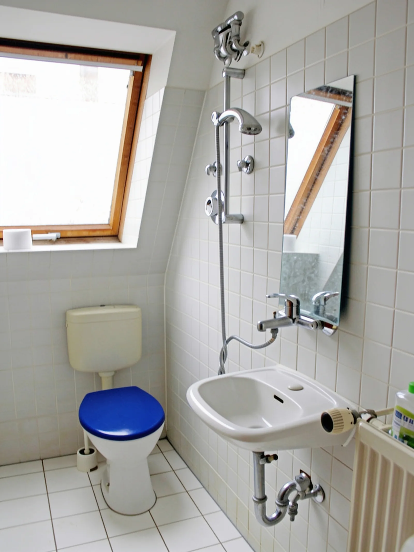 Kleines Bad unter einer Dachschräge vor der Sanierung mit weißen Fliesen, Stand-WC, Wandwaschbecken und Dachfenster