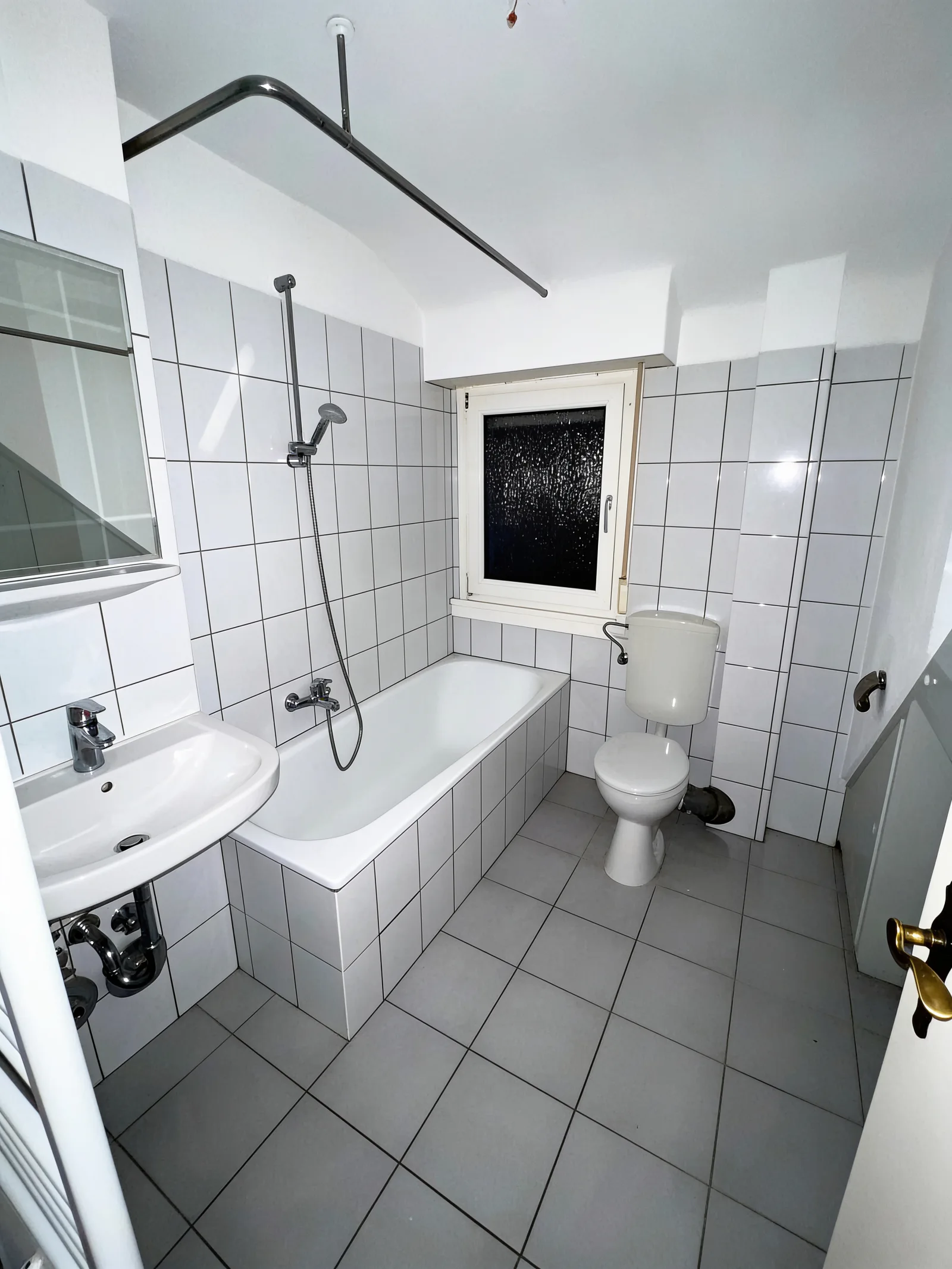 Hell gefliestes Bad vor der Sanierung mit Badewanne, Stand-WC, kleinem Waschbecken und Fenster